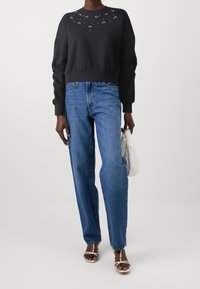 Personne portant un haut noir à manches longues avec de petits motifs blancs, un jean bleu, des sandales blanches à lanières, et tenant un sac matelassé blanc.