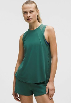 Femeie cu părul prins la spate, purtând un top sportiv verde închis fără mâneci și pantaloni scurți asortați, pe un fundal simplu și deschis la culoare.