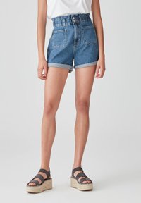 Persona que lleva pantalones cortos de mezclilla azules de talle alto con dobladillos enrollados, combinados con sandalias de plataforma negras y una blusa blanca metida por dentro.