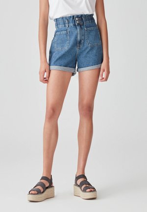 Person wearing high-waisted blue denim shorts with rolled hems, paired with black platform sandals and a white tucked-in top.
