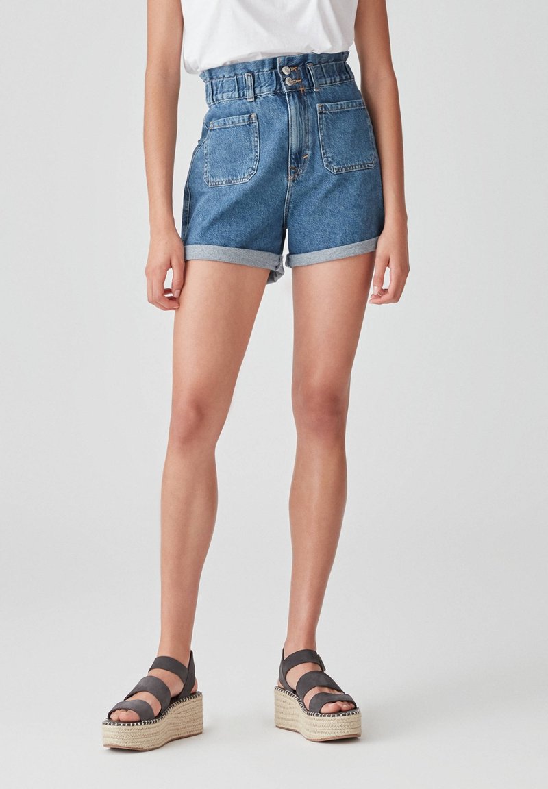 Persona que lleva pantalones cortos de mezclilla azules de talle alto con dobladillos enrollados, combinados con sandalias de plataforma negras y una blusa blanca metida por dentro.