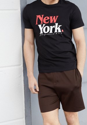 Schwarzes T-Shirt mit der Aufschrift "New York" in Rot und Weiß, kombiniert mit braunen Nadelstreifen-Shorts. Der Stoff wirkt glatt und der Schnitt ist leger.