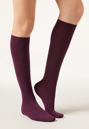 Piernas con calcetas altas hasta la rodilla de color morado oscuro sólido, de pie sobre un fondo claro y liso, con un pie ligeramente levantado.