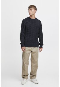 Dunkelblauer Strickpullover mit Kabelmuster, Rundhalsausschnitt und gerippten Bündchen, kombiniert mit beigen straight-leg Hosen und weißen Sneakers.