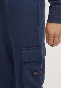 Cargo hlače u tamnoplavom pamučnom materijalu s dva bočna džepa i velikim zakrpa džepom na bedru. Sadrži aplikaciju sa šivenim logotipom.