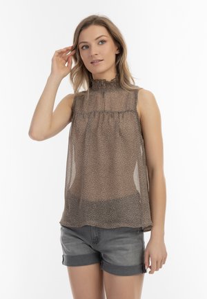 Femme aux cheveux châtain clair portant un chemisier sans manches à motifs et un short en jean gris, touchant ses cheveux et regardant légèrement à droite.