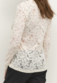 Krämfärgad blus med lång ärm i spets med ett blommönster. Genomskinlig textur med täta vävningar och scallopad nederkant, mot en svart bakgrund.