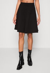 Černá sukně ke kolenům vyrobená z texturované látky, se přiléhavým pasem a rozšířeným siluetou. Spárována s černými kotníkovými botami.