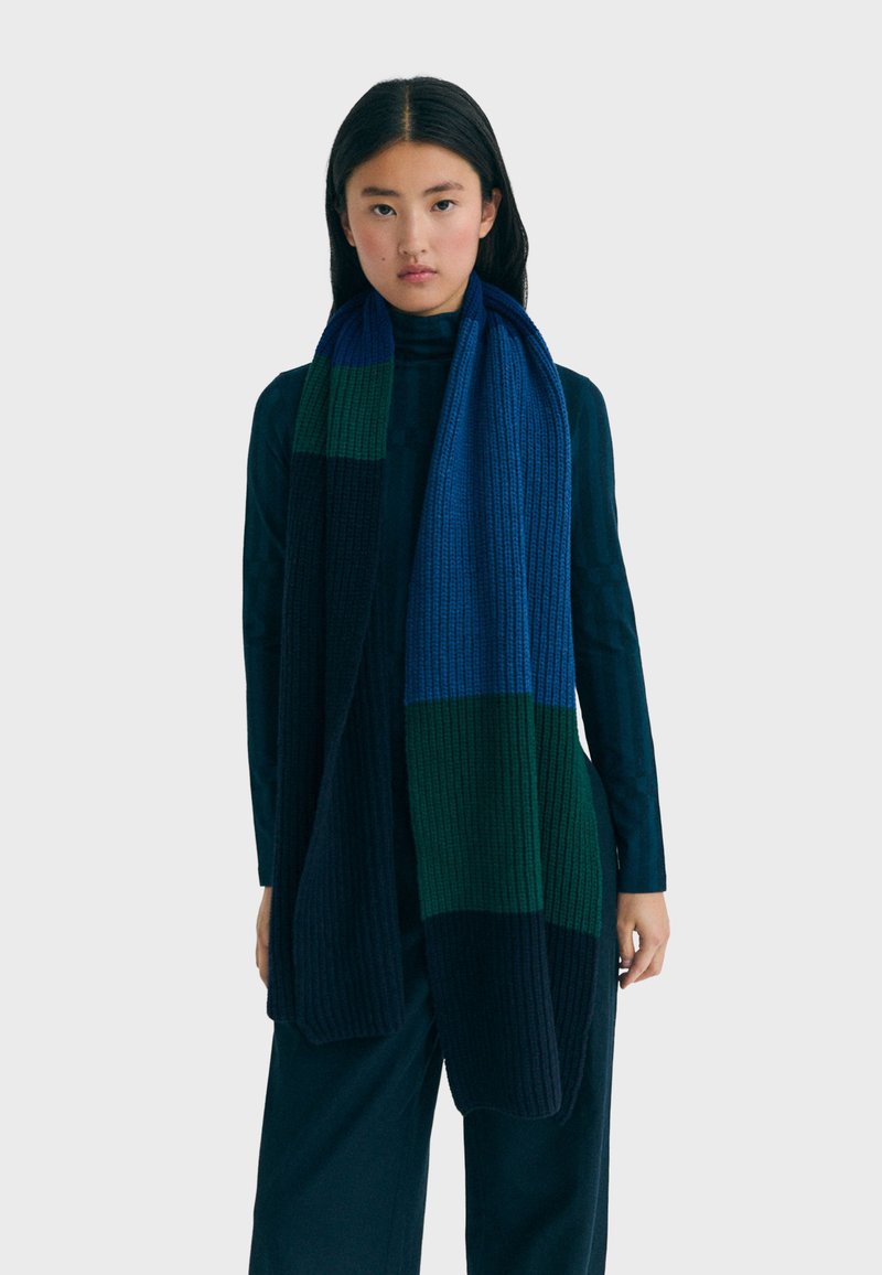 Écharpe tricotée avec un design en blocs de couleurs bleu, vert et bleu marine foncé. La texture côtelée offre une sensation de chaleur et de douceur avec une forme longue et tombante.