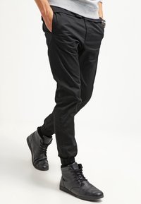 Svarta tapered byxor med elastiska manschetter, tillverkade av slät tyg. Bärs med mörka hög-top sneakers som har snörningsdesign och strukturerade detaljer.