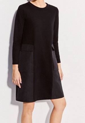 Czarna długa sukienka z długim rękawem sięgająca kolan, z okrągłym dekoltem i dwoma dużymi przednimi kieszeniami, noszona przez osobę na jednolitym tle.