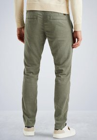 Pantalones ajustados de color verde caqui, confeccionados en una tela suave y texturizada, con dos bolsillos traseros, costuras alineadas y combinados con zapatos blancos.