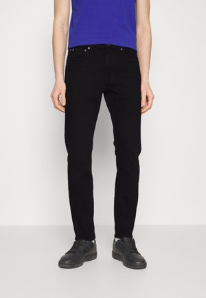 Man draagt zwarte slim-fit jeans, zwarte sneakers en een blauw overhemd, staand tegen een effen witte achtergrond.