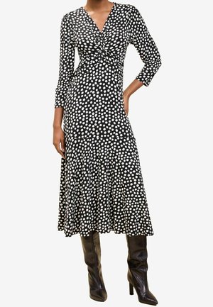 Femme portant une robe midi noire à pois blancs, manches trois-quarts et bottes noires à talons jusqu'au genou, debout avec une main sur la hanche.