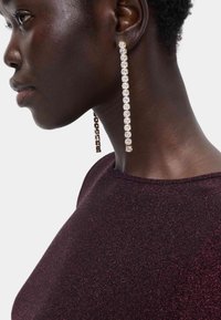 Długie, wiszące kolczyki z przezroczystymi kryształami osadzonymi w złotym odcieniu, zwisające z geometrycznego łańcucha, noszone przez osobę w ciemnopurpurowej bluzce.