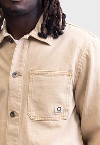 Chaqueta beige con cierre de botones en la parte delantera, bolsillo en el pecho izquierdo y parche del logo. La tela parece robusta con detalles de costura visibles.