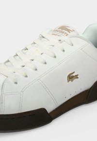 Λευκό δερμάτινο sneaker Lacoste με λευκά κορδόνια, μπεζ λογότυπο κροκόδειλου στο πλάι, μπεζ σήμανση στη γλώσσα και μαύρη-καφέ σόλα.