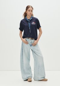 Marineblå bomulds-t-shirt med optrukne ærmer, med "BRUNCH SPOT" grafik. Lyst vaskede vide bukser; beige sko uden snørebånd. Blå halsklud.