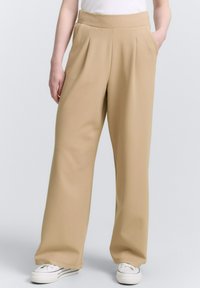 Pantaloni beige a gamba larga con una texture liscia, frontale plissettato e tasche laterali, abbinati a sneakers bianche per un contrasto.