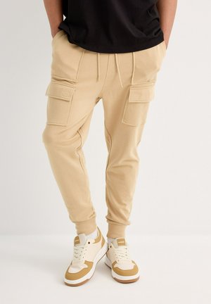 Pantalones cargo beige con múltiples bolsillos laterales, cintura con cordón y tobillos ajustados, combinados con zapatillas blancas y beiges.