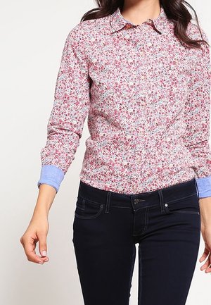 Kvinde iført en langærmet, knappet skjorte med et lille blomsterprint og mørkeblå, tætsiddende jeans.