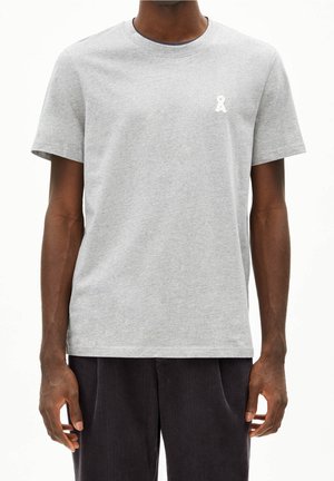 Mężczyzna noszący jasnoszary t-shirt z krótkim rękawem z małym białym logo na lewej piersi, w połączeniu z ciemnymi spodniami.