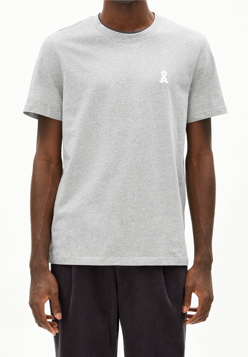 Homme portant un t-shirt gris clair à manches courtes avec un petit logo blanc sur le côté gauche de la poitrine, associé à un pantalon foncé.