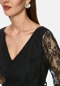 Schwarzes Spitzenkleid mit V-Ausschnitt, langen Spitzenärmeln und einem strukturierten Oberteil. Goldene, runde Ohrringe betonen den Look.