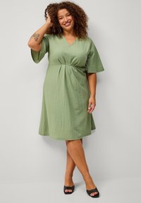 Grünes knielanges Kleid aus strukturiertem Stoff mit V-Ausschnitt und kurzen Glockenärmeln; mit gerafftem Taillendetail. Paar schwarze High Heels.