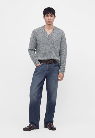 Homme portant un pull en maille gris à col en V, un jean droit bleu foncé avec une ceinture marron, et des mocassins marron foncé, debout devant un fond blanc.