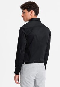 Maglietta nera a maniche lunghe con colletto strutturato, texture liscia, patta dei bottoni nascosta e vestibilità su misura, abbinata a pantaloni a righe check grigio chiaro.