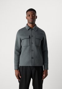 Gray wool shirt jacket with a collar, two chest pockets, front button closure, and long sleeves, paired with black pants.
