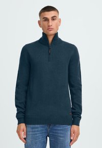 Dunkeltürkiser Pullover mit hohem Kragen, halbem Reißverschluss, gerippter Textur und enganliegenden Manschetten. Über blauen Jeans getragen. Etikett unten sichtbar.