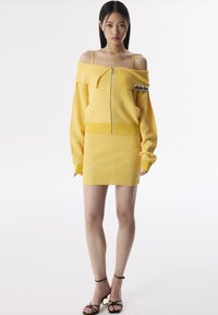 Pull jaune en tricot décolleté avec fermeture éclair à l'avant et détail de logo, associé à une jupe mini texturée assortie et des sandales noires à lanières.