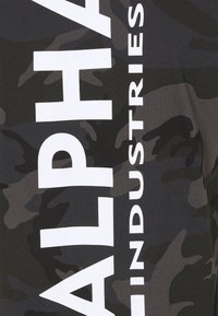 Svart T-skjorte med camouflage-mønster og stor hvit tekst som leser "ALPHA INDUSTRIES" vertikalt på venstre side. Laget av bomull.