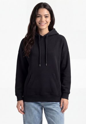 Ung kvinde med langt mørkt hår iført en sort hættetrøje og lyseblå jeans, smilende og stående foran en ensfarvet hvid baggrund.