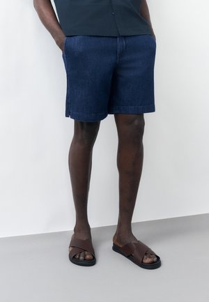 Mężczyzna w ciemnoniebieskich szortach dżinsowych, czarnej koszuli i brązowych skórzanych sandałach stoi przy białej ścianie, ręce w kieszeniach.