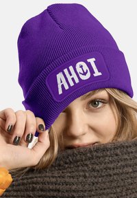 Purple knitted beanie featuring an embroidered "AHOI" patch with an anchor design. The fabric has a ribbed texture and a snug fit.