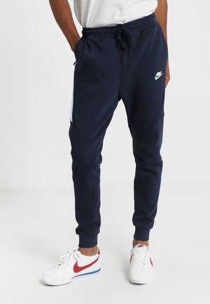 Mann trägt marineblaue Nike-Jogginghosen mit weißen und roten Nike-Sneakers, Hände in den Taschen, stehend vor einfachem weißem Hintergrund.