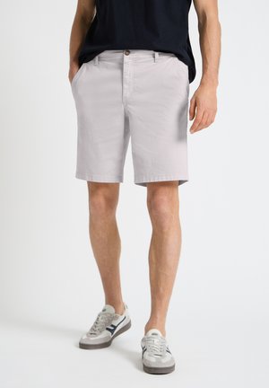 Mann trägt hellgraue knielange Shorts, schwarzes Shirt und weiße Turnschuhe mit dunklen Akzenten, steht mit den Händen in den Taschen.