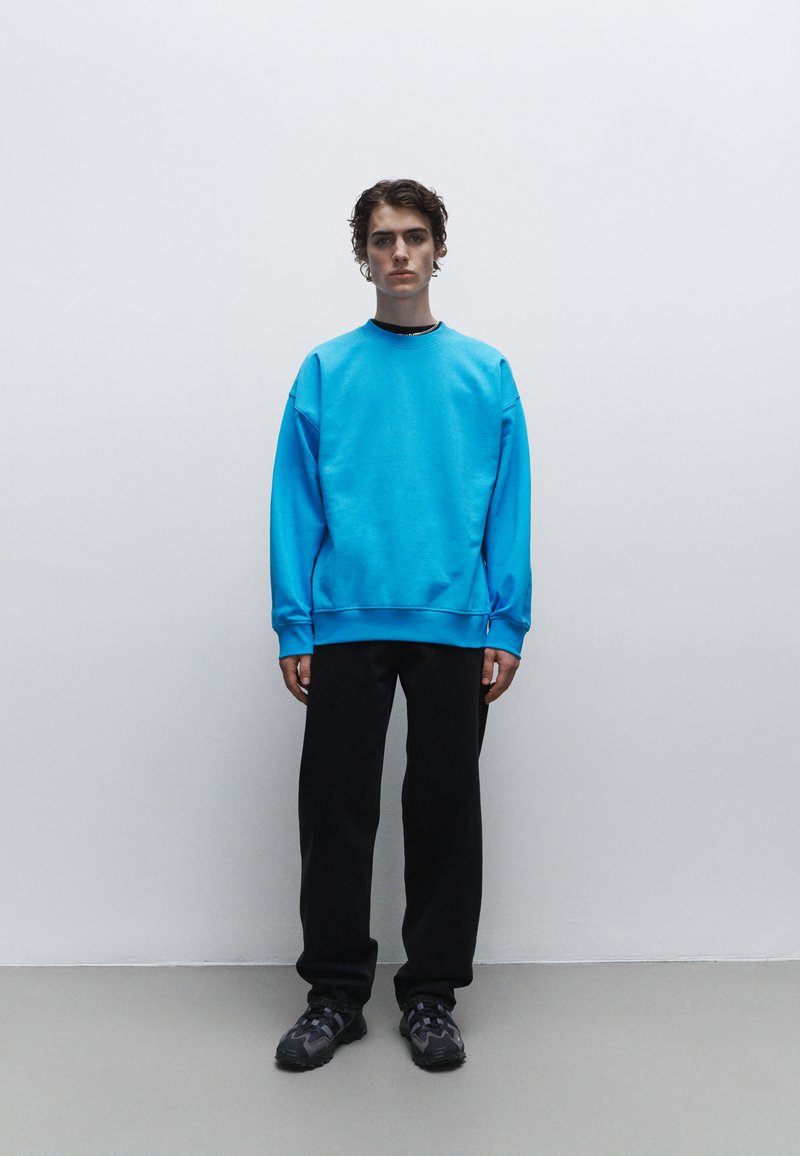Blauwe sweatshirt met verlaagde schouders, ribgebreide boorden en zoom; gedragen met zwarte broek en gestructureerde zwarte schoenen. Effen achtergrond.