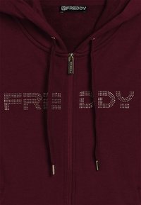 Felpa con zip color mogano, dotata di lacci e borchie metalliche che formano la scritta "FREDDY" sul petto, sotto un'etichetta con il marchio.