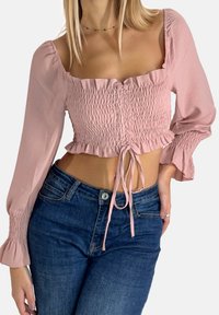 Pink croppet top med smocket tekstur og flæse detaljer; har lange ærmer og bundet front, kombineret med mørke denim jeans.