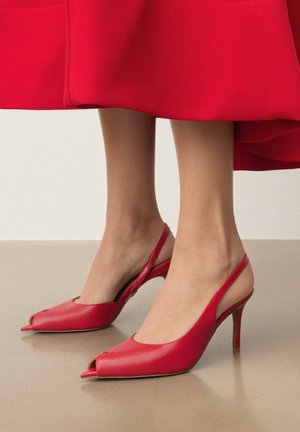 Lábak piros bőrből készült bokapántos, nyitott orrú magas sarkú szandált viselnek, amelyet piros szoknyával párosítanak semleges padló és háttér előtt.