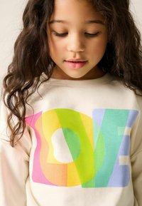 Krémová mikina má velký grafický prvek "LOVE" v překrývajících se pastelových barvách: růžové, oranžové, žluté, zelené a modré s jemnou strukturou.