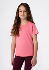 Fiatal lány, hosszú göndör hajjal, rózsaszín, rövid ujjú pólót és sötét nadrágot visel, egy sima világos háttér előtt áll.