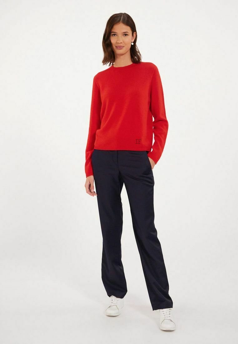 Pull en tricot rouge avec un col rond, associé à un pantalon marine et des baskets blanches. Le pull présente un détail logo subtil à l'ourlet.