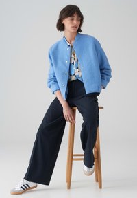 Bomber jacket in lana azzurro chiaro con bottoni a pressione argento, abbinato a una maglietta a fantasia, pantaloni neri e sneakers bianche con accenti grigi.