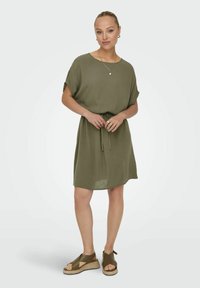 Olivgrünes, kurzärmeliges Kleid mit rundem Ausschnitt und einem Bindeband in der Taille; kombiniert mit olivgrünen Espadrilles.