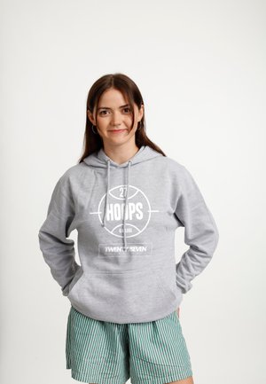 Ung kvinde med brunt hår iført grå "Hoops" hættetrøje og grønstribede shorts, står med hænderne i lommerne mod en hvid baggrund.